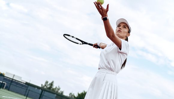 Attractive woman in white sportswear with the racket and the ball in her hands is ready to hit the ball on the court